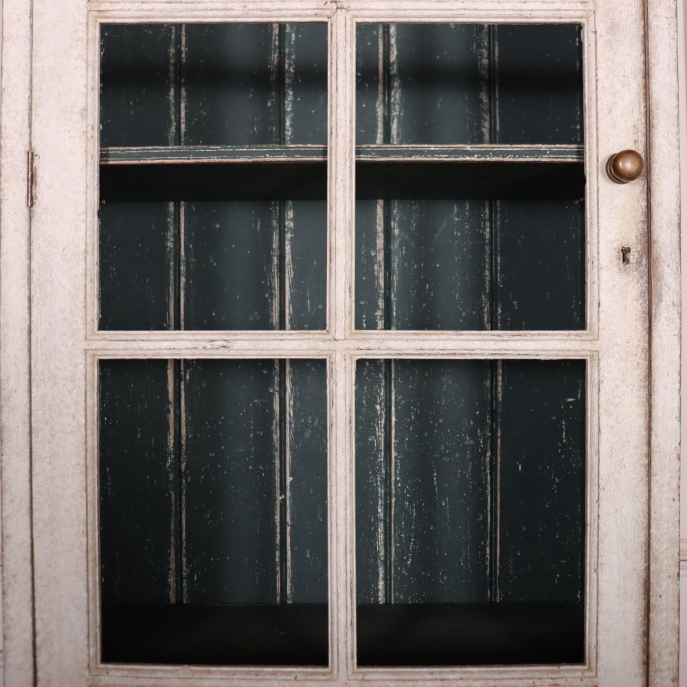 A tall, narrow off-white painted glazed bookcase featuring a glass upper section with a dark interior, above a single panelled cupboard door. View IV