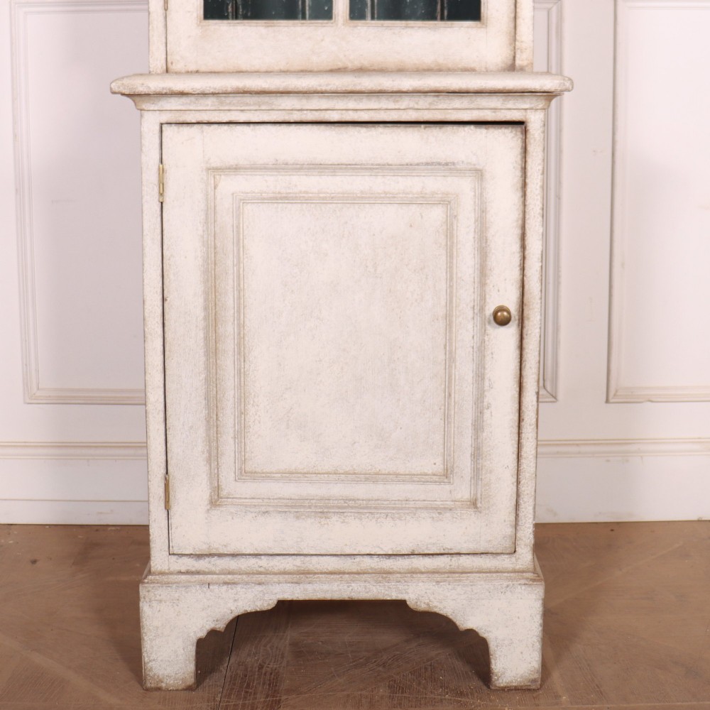 A tall, narrow off-white painted glazed bookcase featuring a glass upper section with a dark interior, above a single panelled cupboard door. View V