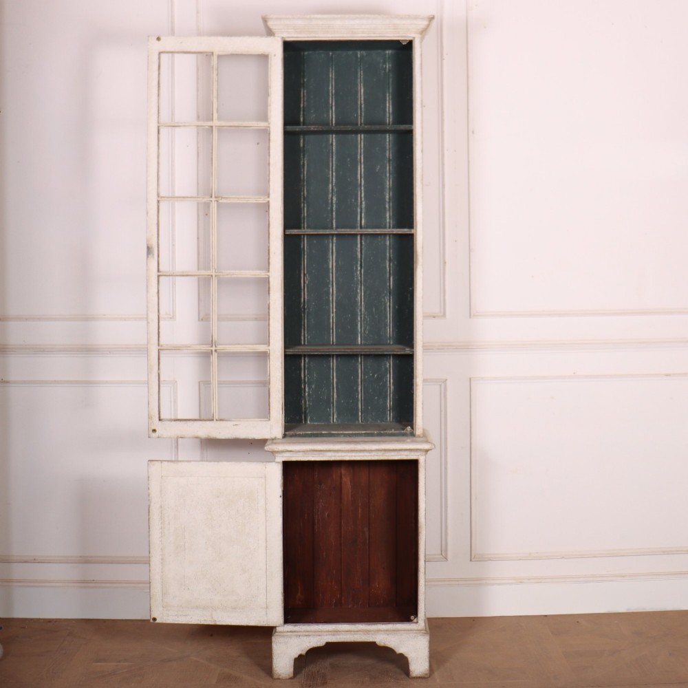 A tall, narrow off-white painted glazed bookcase featuring a glass upper section with a dark interior, above a single panelled cupboard door. View VI