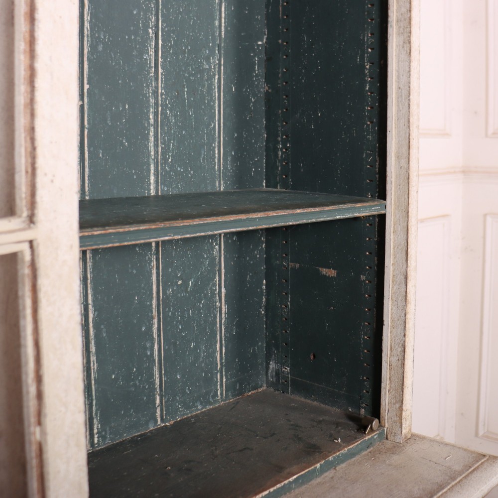 A tall, narrow off-white painted glazed bookcase featuring a glass upper section with a dark interior, above a single panelled cupboard door. View VII