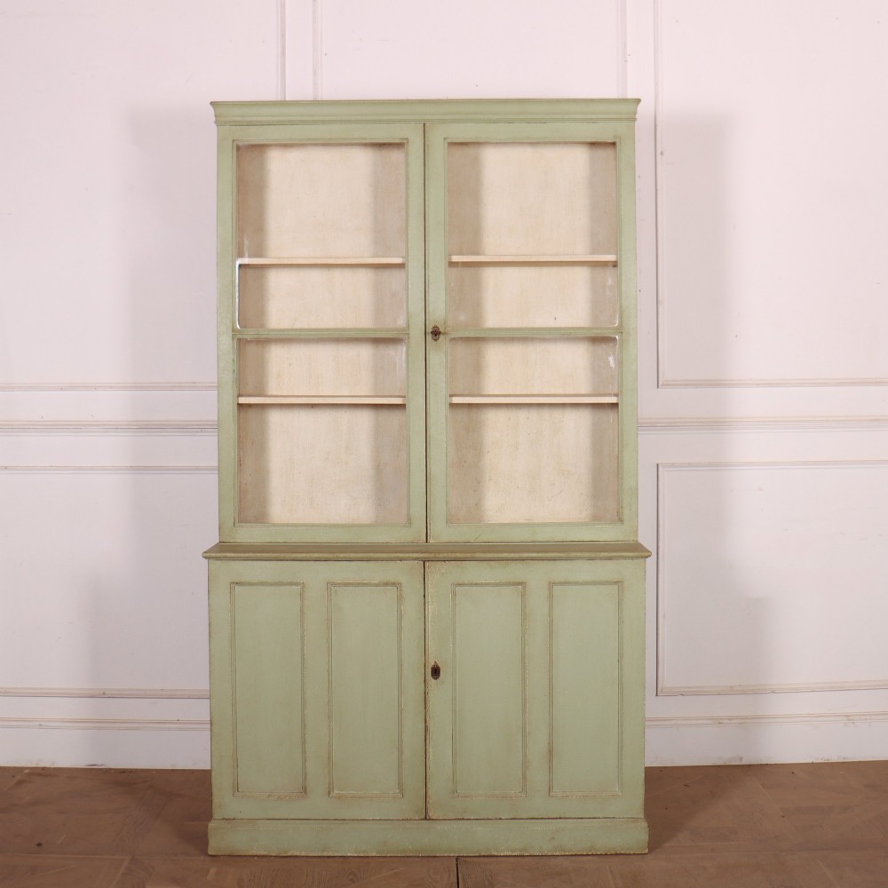 A tall sage green painted bookcase featuring two upper glazed doors revealing interior shelving, resting upon a base cabinet with two panelled doors.