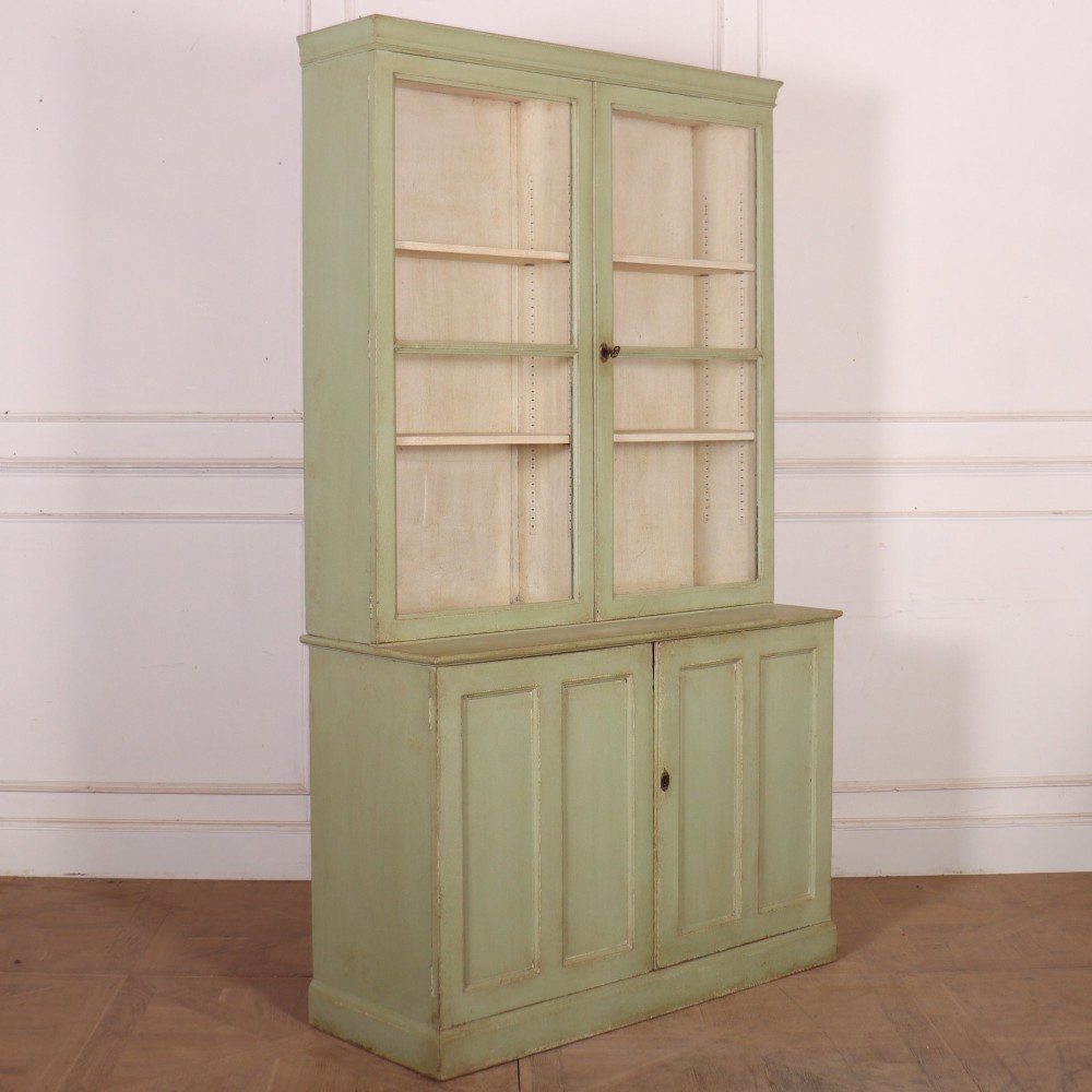 A tall sage green painted bookcase featuring two upper glazed doors revealing interior shelving, resting upon a base cabinet with two panelled doors. View X
