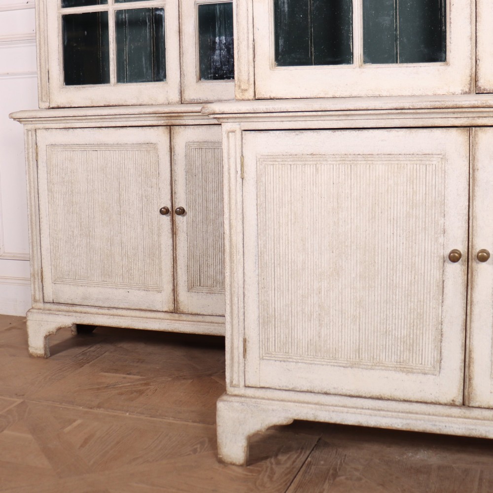 A pair of tall Swedish Gustavian style off-white painted bookcases featuring glazed upper doors with dark green interior backs above lower cupboards. View VI