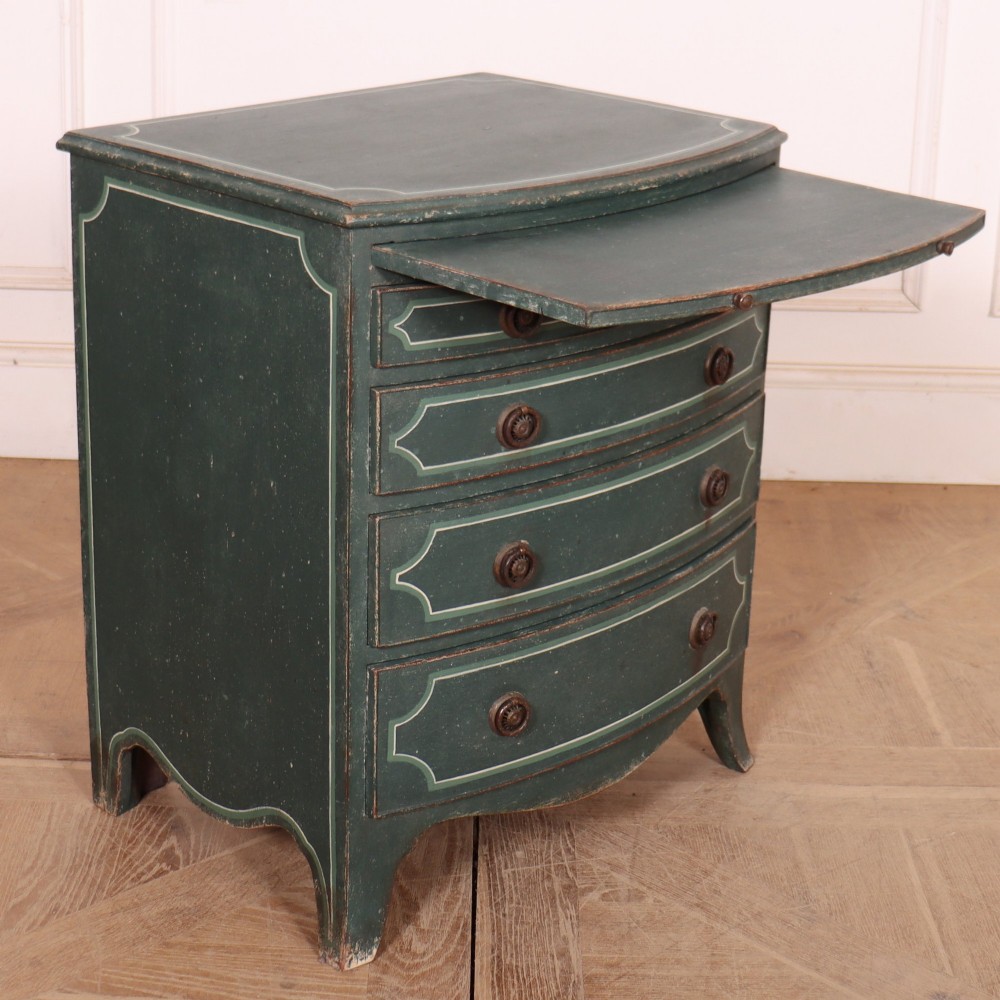 A deep green painted bowfront bedside chest of drawers featuring light green decorative panel outline details, brass ring handles, and splayed legs. View VII