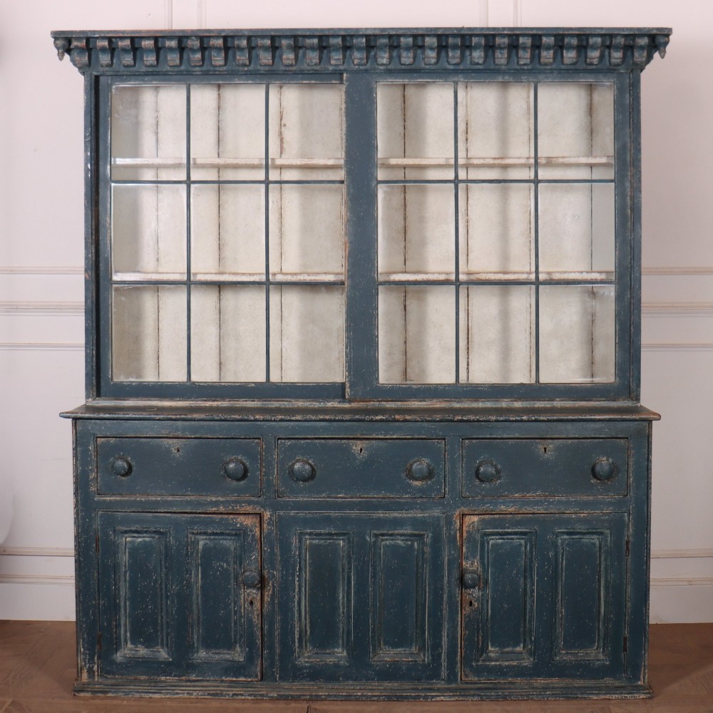 A large two-part dark blue painted timber dresser featuring a glazed upper display section with grid-paned doors and a dentil corniced top, resting on a base with three drawers and three panelled cupboard doors.
