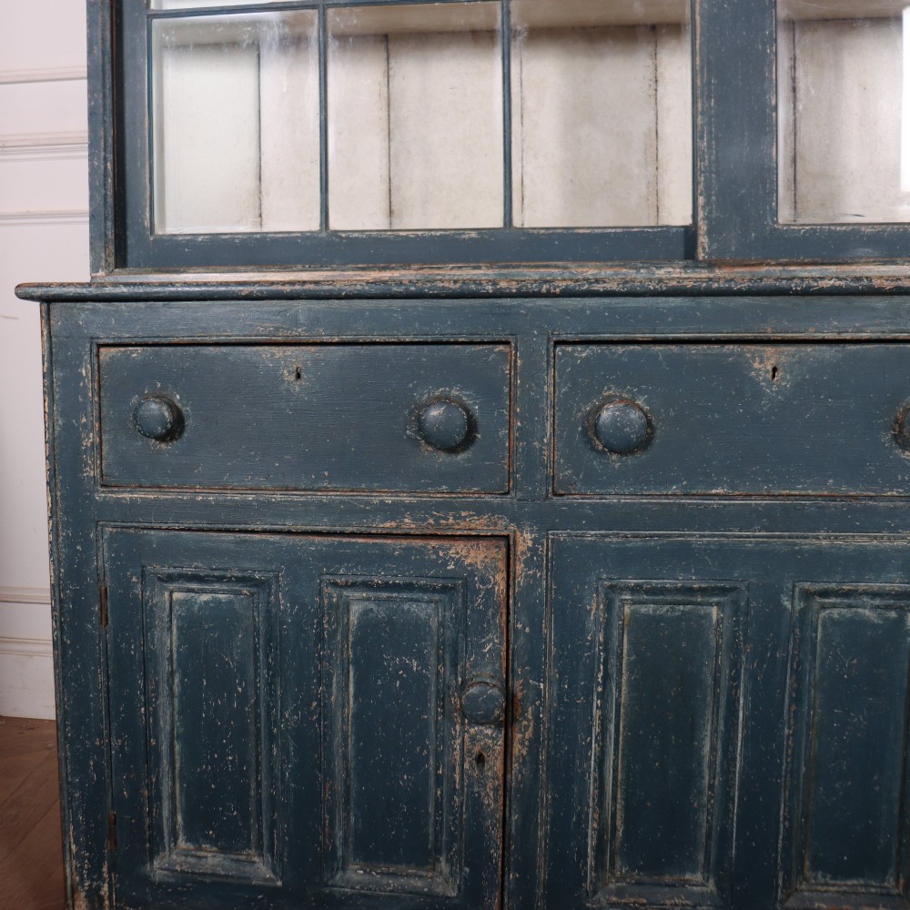 A large two-part dark blue painted timber dresser featuring a glazed upper display section with grid-paned doors and a dentil corniced top, resting on a base with three drawers and three panelled cupboard doors. View II