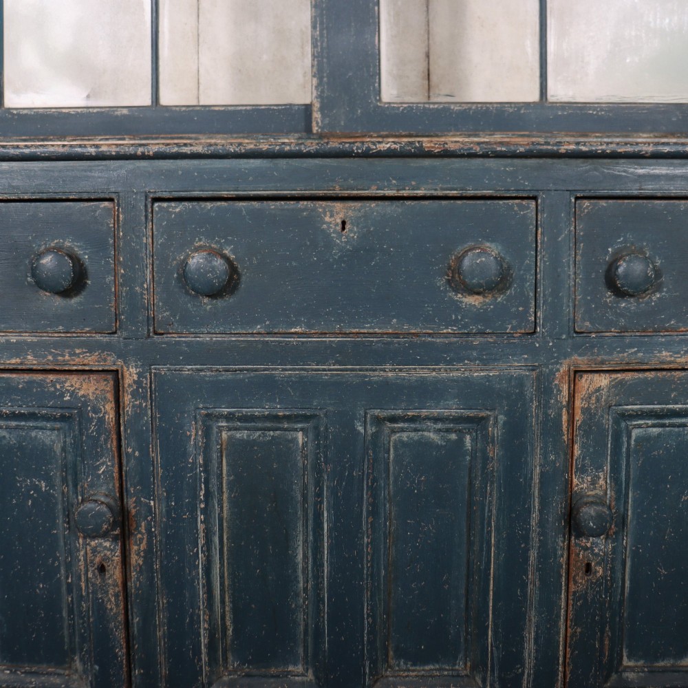 A large two-part dark blue painted timber dresser featuring a glazed upper display section with grid-paned doors and a dentil corniced top, resting on a base with three drawers and three panelled cupboard doors. View IV