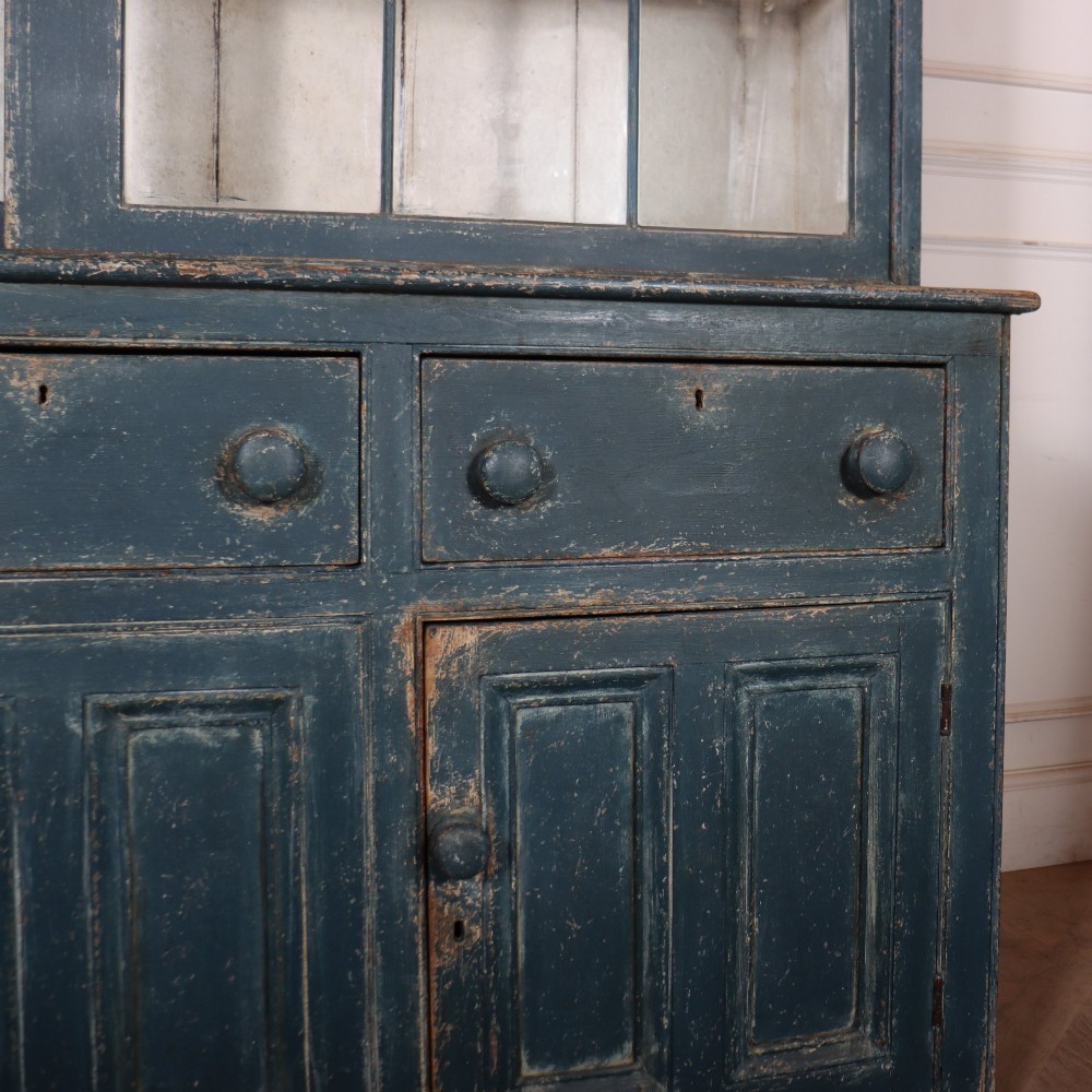 A large two-part dark blue painted timber dresser featuring a glazed upper display section with grid-paned doors and a dentil corniced top, resting on a base with three drawers and three panelled cupboard doors. View VI