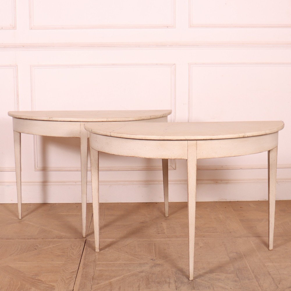 A pair of off-white painted semicircular demi lune console tables with slender tapered legs resting on a wooden parquet floor.