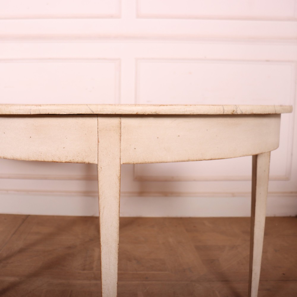 A pair of off-white painted semicircular demi lune console tables with slender tapered legs resting on a wooden parquet floor. View II