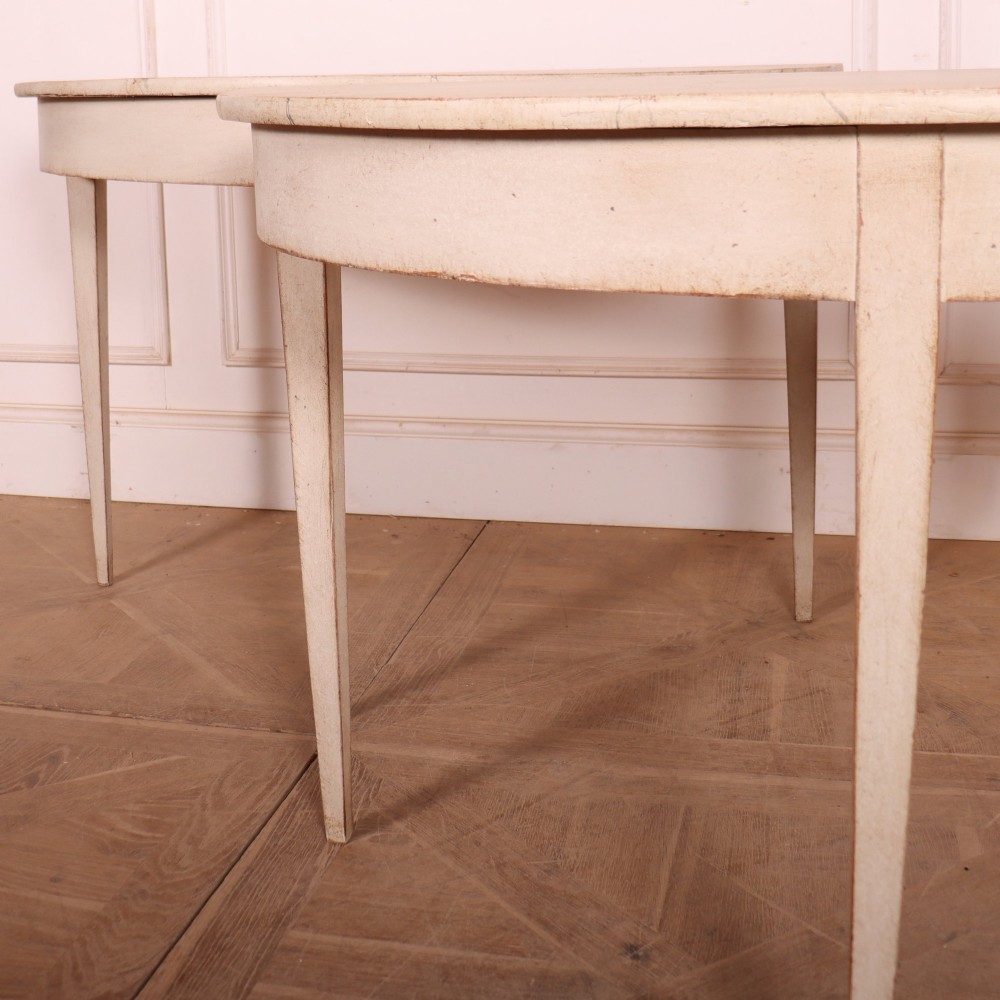 A pair of off-white painted semicircular demi lune console tables with slender tapered legs resting on a wooden parquet floor. View III