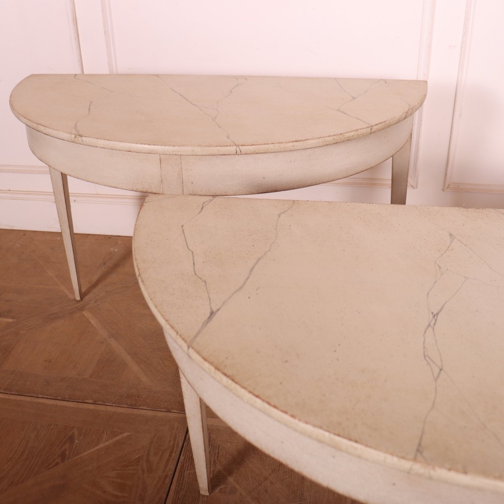 A pair of off-white painted semicircular demi lune console tables with slender tapered legs resting on a wooden parquet floor. View IV
