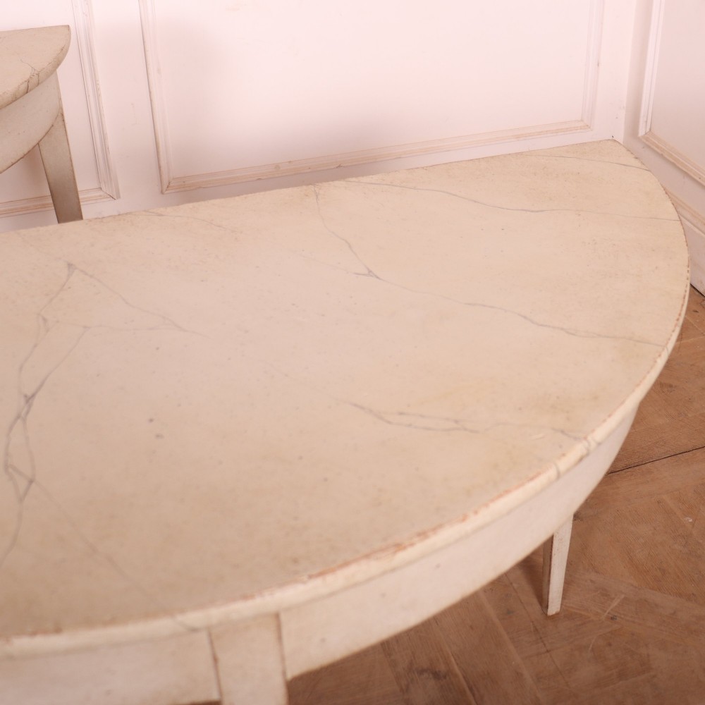 A pair of off-white painted semicircular demi lune console tables with slender tapered legs resting on a wooden parquet floor. View V