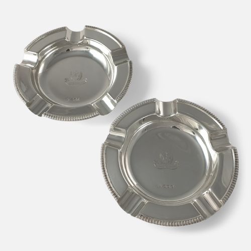 fig 1. A pair of round sterling silver ashtrays featuring engraved central crests, visible hallmarks, and gadrooned rims with cigarette rests.