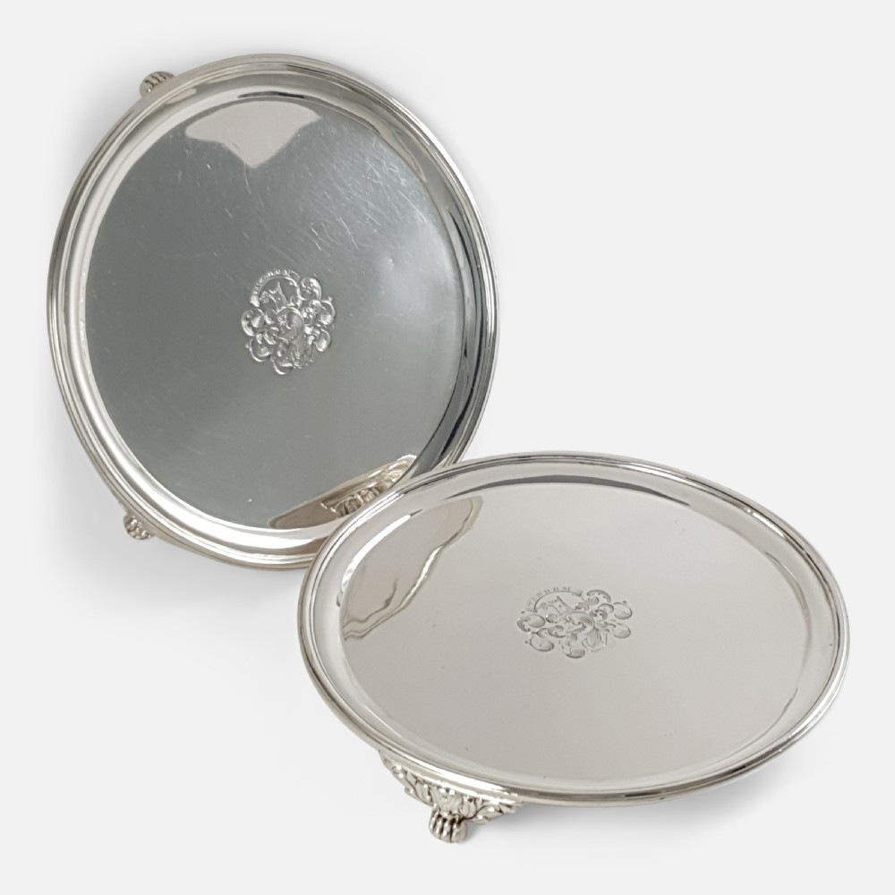 A pair of circular 1818 George III sterling silver salvers featuring central engraved crests, moulded rims, and short bracket feet.