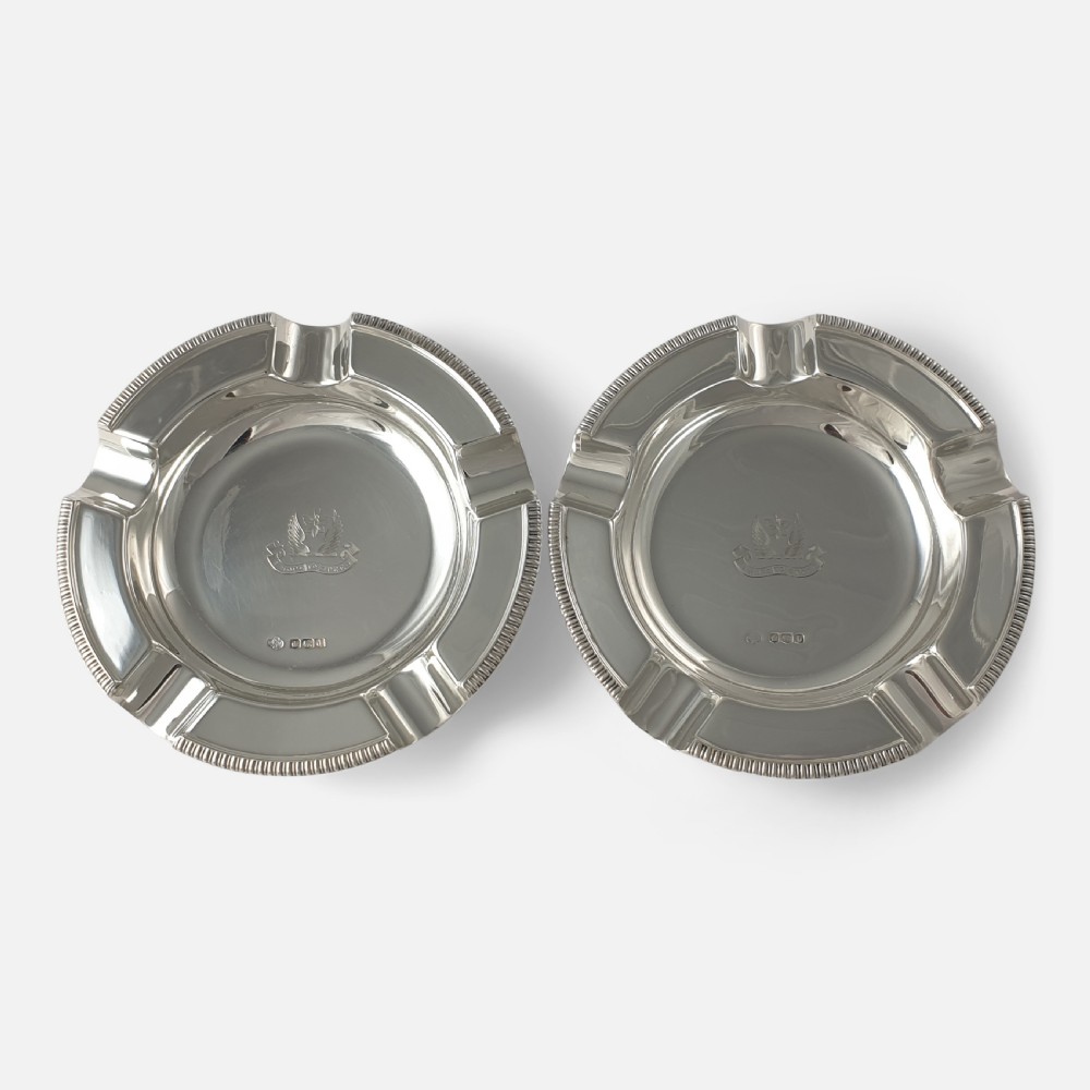 A pair of round sterling silver ashtrays featuring engraved central crests, visible hallmarks, and gadrooned rims with cigarette rests.