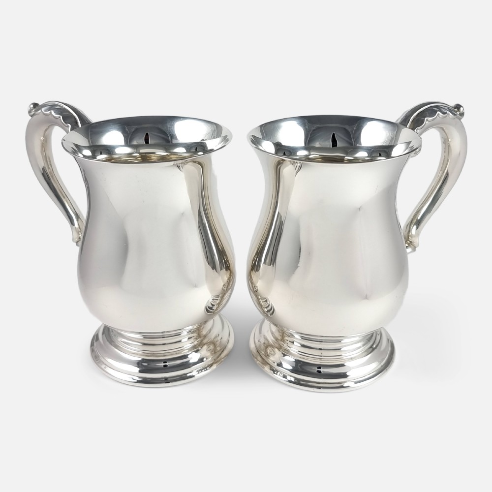 A pair of polished sterling silver mugs featuring baluster-shaped bodies, stepped circular pedestal bases, and curved scroll handles.