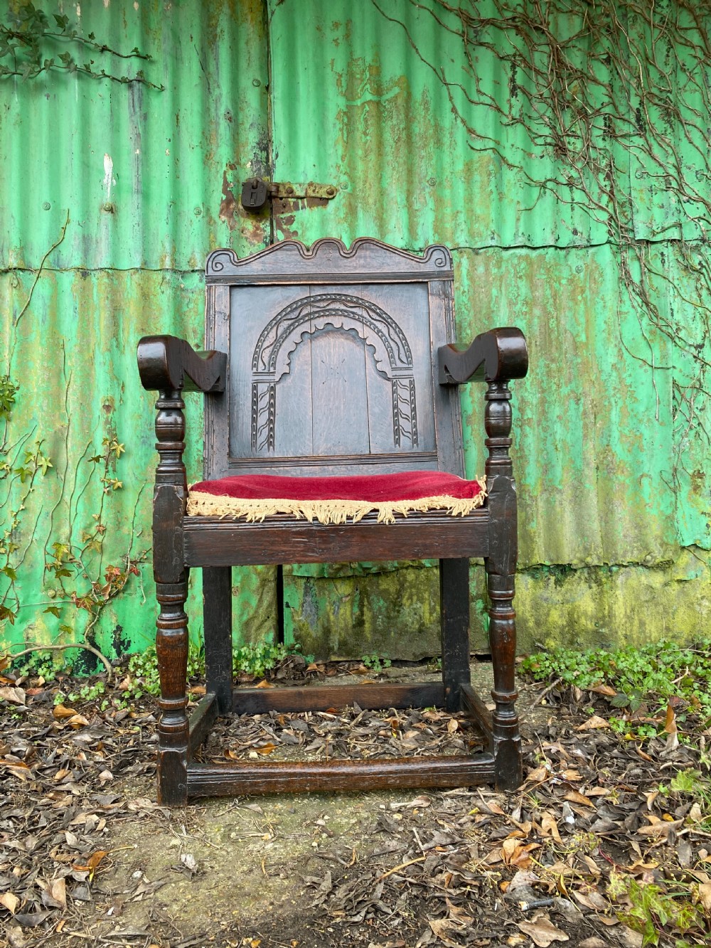Carved dark oak wainscot armchair featuring an arched back panel, turned front supports, and a red velvet seat cushion with fringe trim.