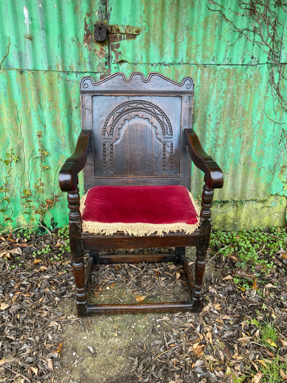 Carved dark oak wainscot armchair featuring an arched back panel, turned front supports, and a red velvet seat cushion with fringe trim. View III