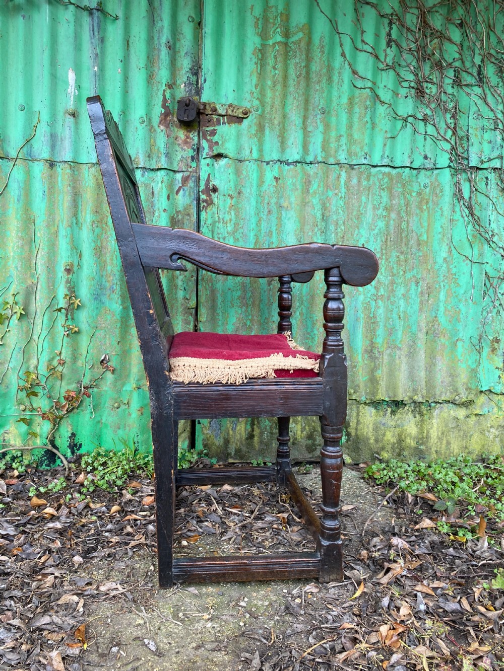Carved dark oak wainscot armchair featuring an arched back panel, turned front supports, and a red velvet seat cushion with fringe trim. View V