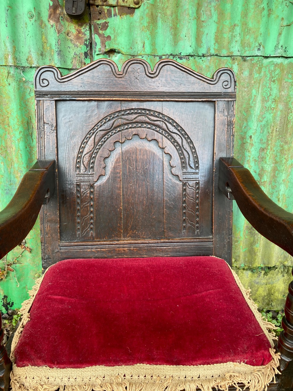 Carved dark oak wainscot armchair featuring an arched back panel, turned front supports, and a red velvet seat cushion with fringe trim. View VII