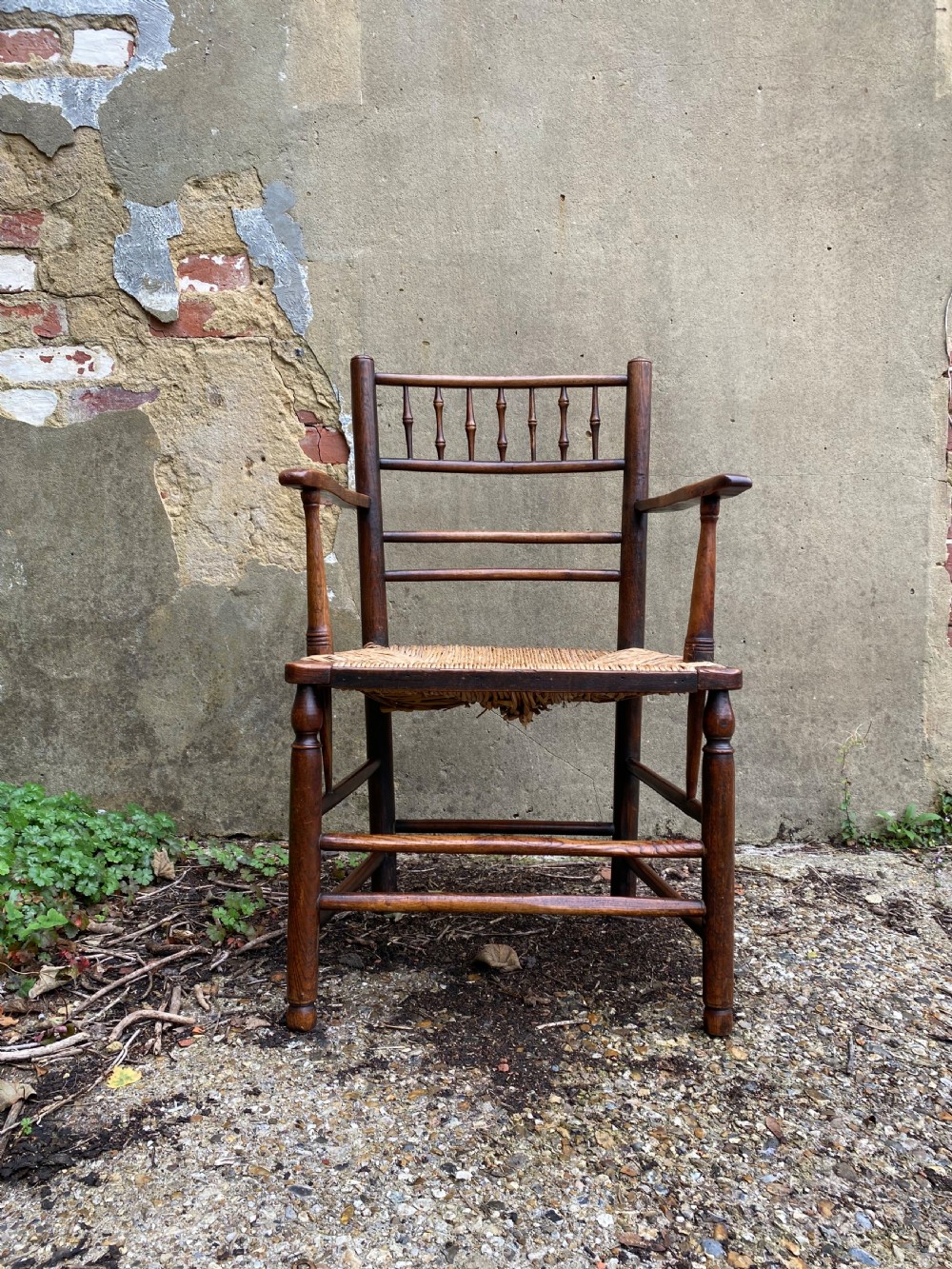 A dark wood Arts and Crafts Sussex armchair featuring a turned spindle backrest, curved armrests, and a woven rush seat.