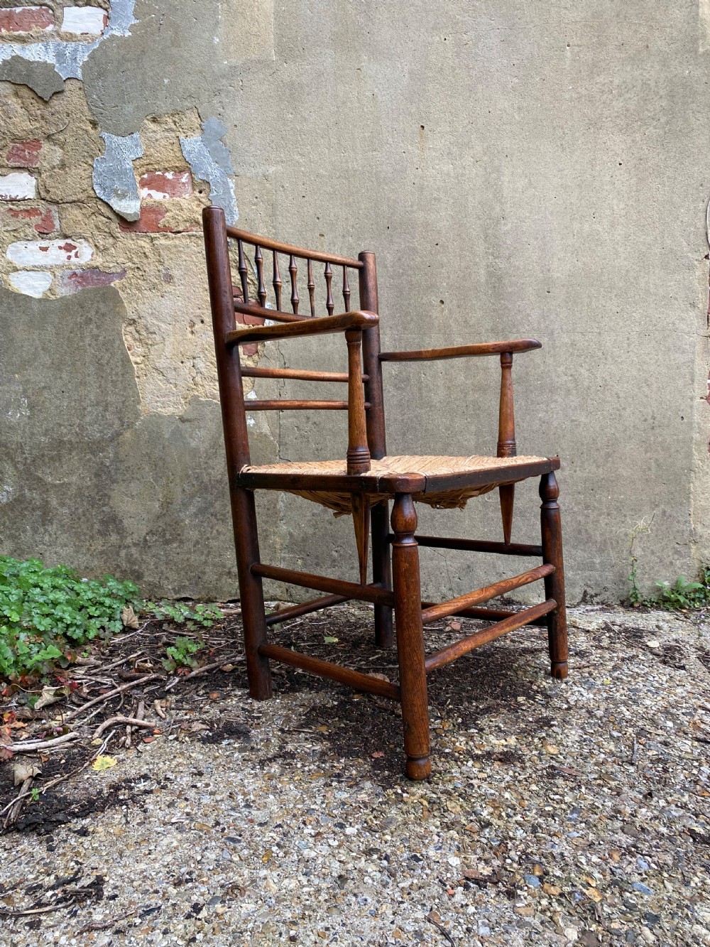 A dark wood Arts and Crafts Sussex armchair featuring a turned spindle backrest, curved armrests, and a woven rush seat. View II