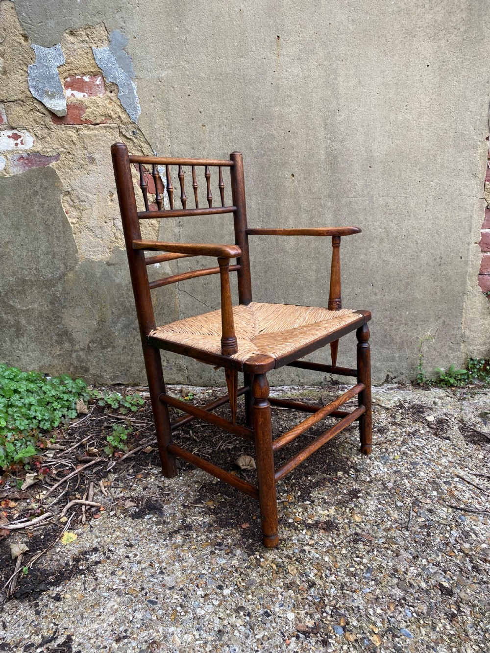 A dark wood Arts and Crafts Sussex armchair featuring a turned spindle backrest, curved armrests, and a woven rush seat. View III