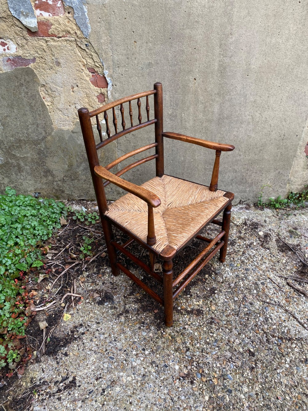 A dark wood Arts and Crafts Sussex armchair featuring a turned spindle backrest, curved armrests, and a woven rush seat. View IV