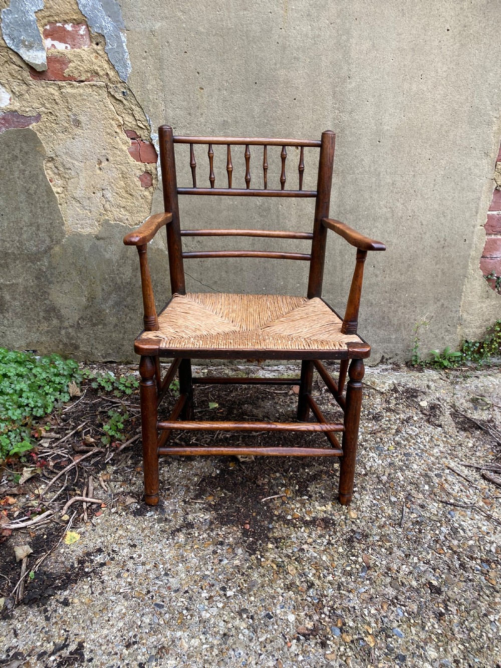A dark wood Arts and Crafts Sussex armchair featuring a turned spindle backrest, curved armrests, and a woven rush seat. View V