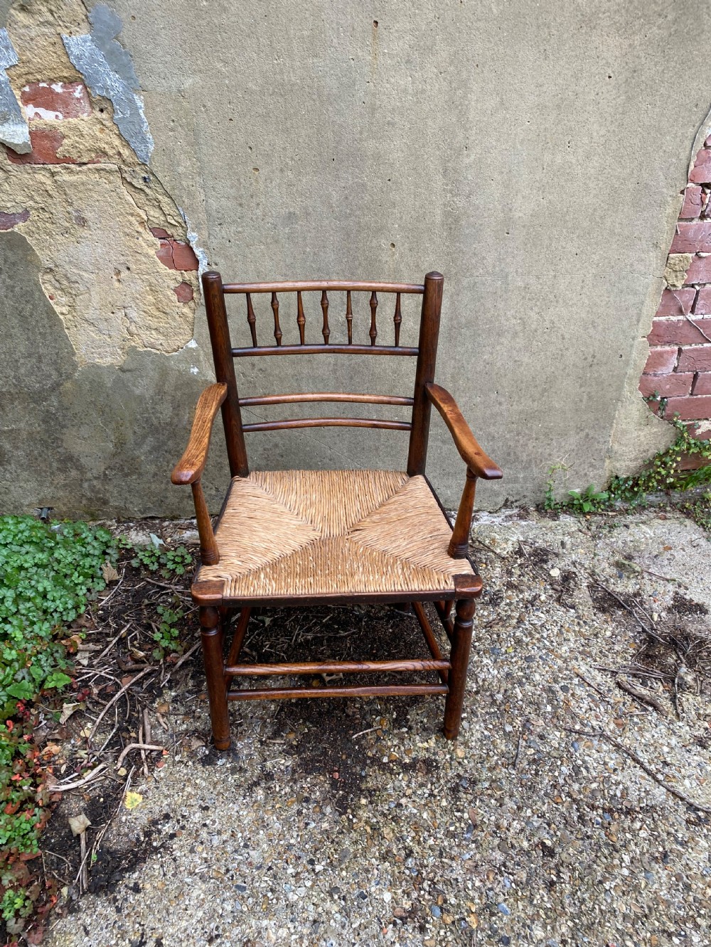 A dark wood Arts and Crafts Sussex armchair featuring a turned spindle backrest, curved armrests, and a woven rush seat. View VI