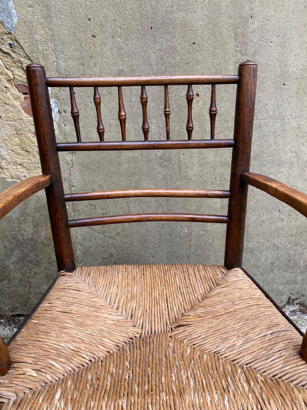 A dark wood Arts and Crafts Sussex armchair featuring a turned spindle backrest, curved armrests, and a woven rush seat. View VII