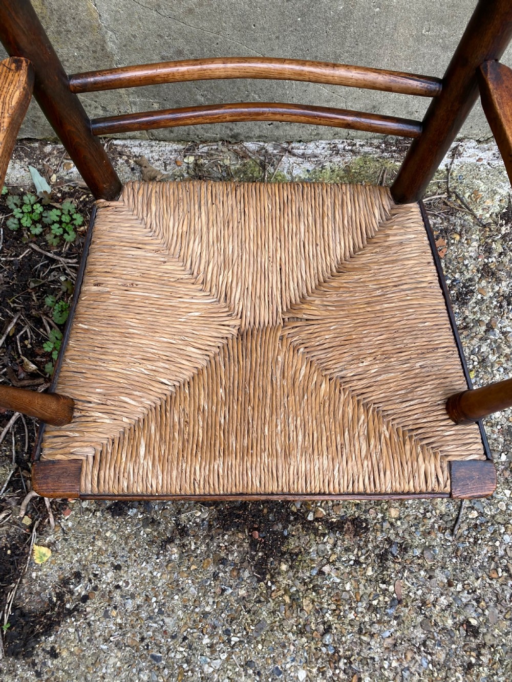 A dark wood Arts and Crafts Sussex armchair featuring a turned spindle backrest, curved armrests, and a woven rush seat. View IX