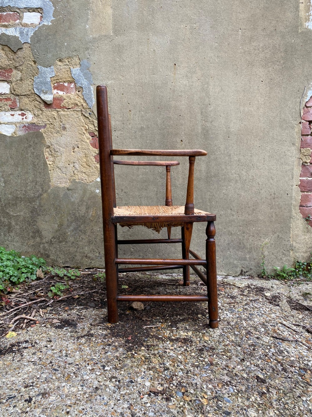 A dark wood Arts and Crafts Sussex armchair featuring a turned spindle backrest, curved armrests, and a woven rush seat. View X