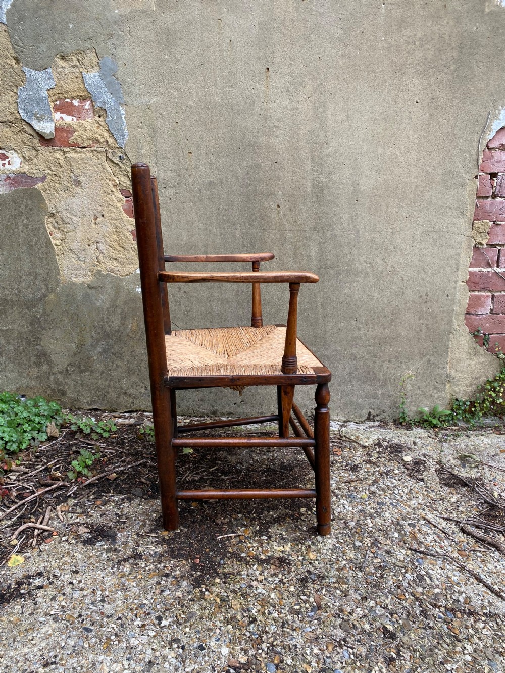 A dark wood Arts and Crafts Sussex armchair featuring a turned spindle backrest, curved armrests, and a woven rush seat. View XI