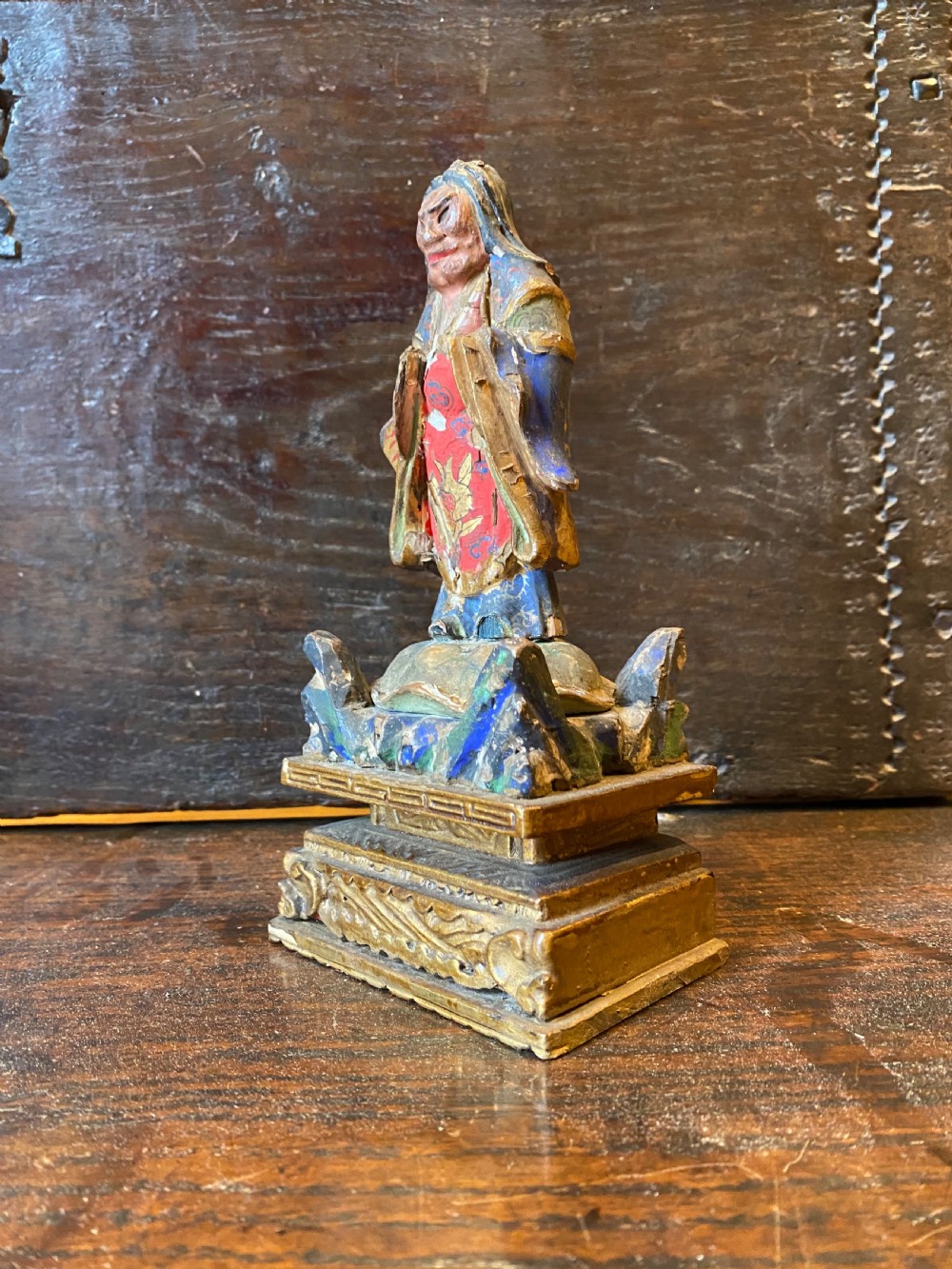 A small hand-carved wooden deity figure with polychrome painted robes and gilt detailing, standing on a carved tiered base. View III