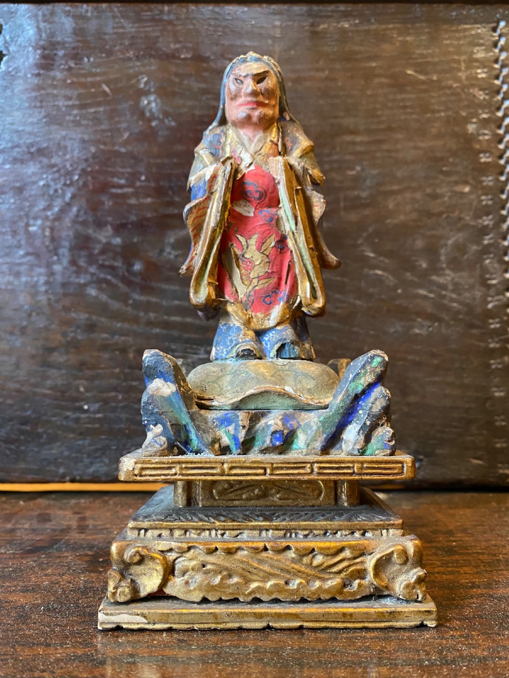A small hand-carved wooden deity figure with polychrome painted robes and gilt detailing, standing on a carved tiered base. View VI