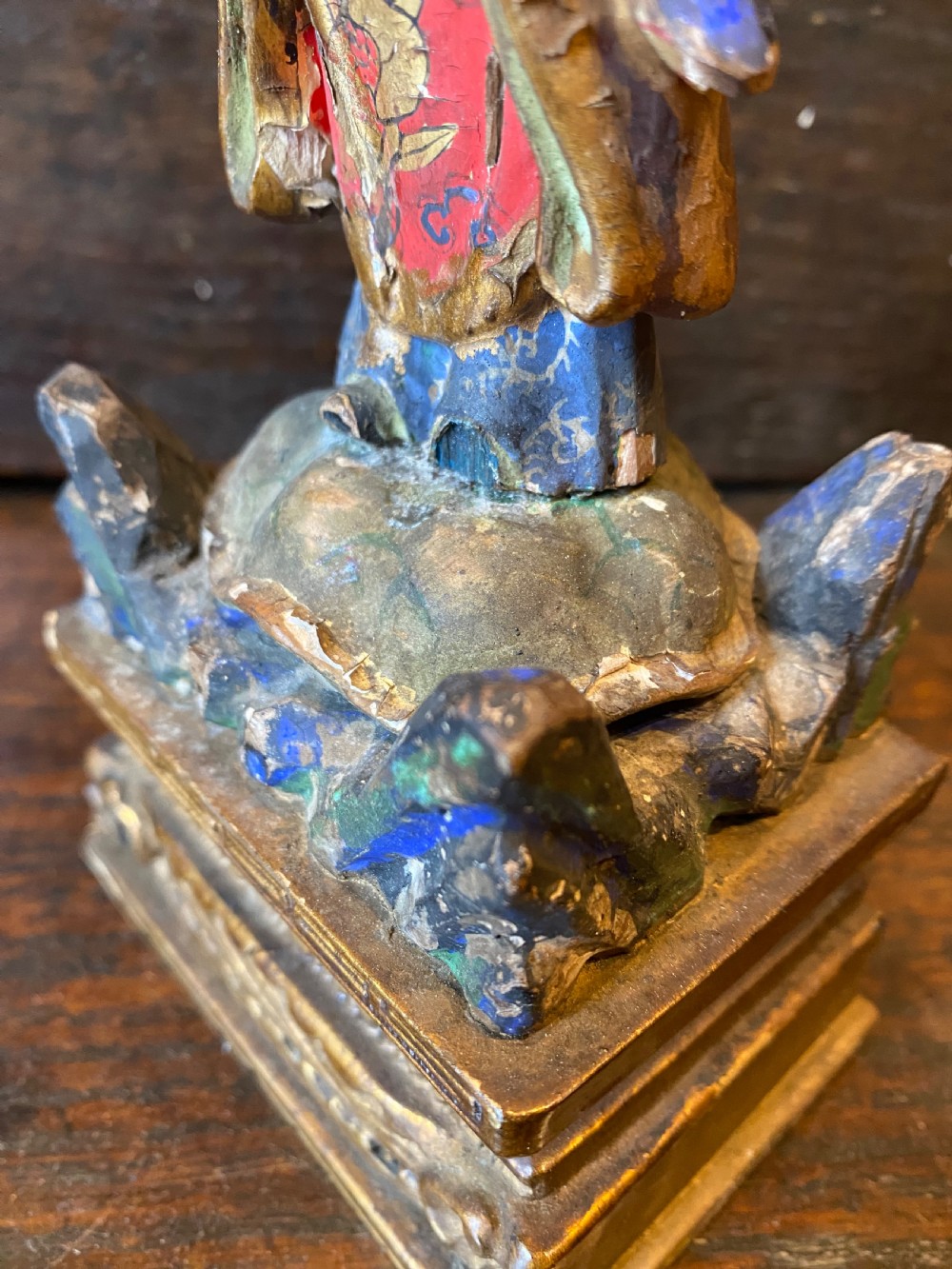 A small hand-carved wooden deity figure with polychrome painted robes and gilt detailing, standing on a carved tiered base. View VII