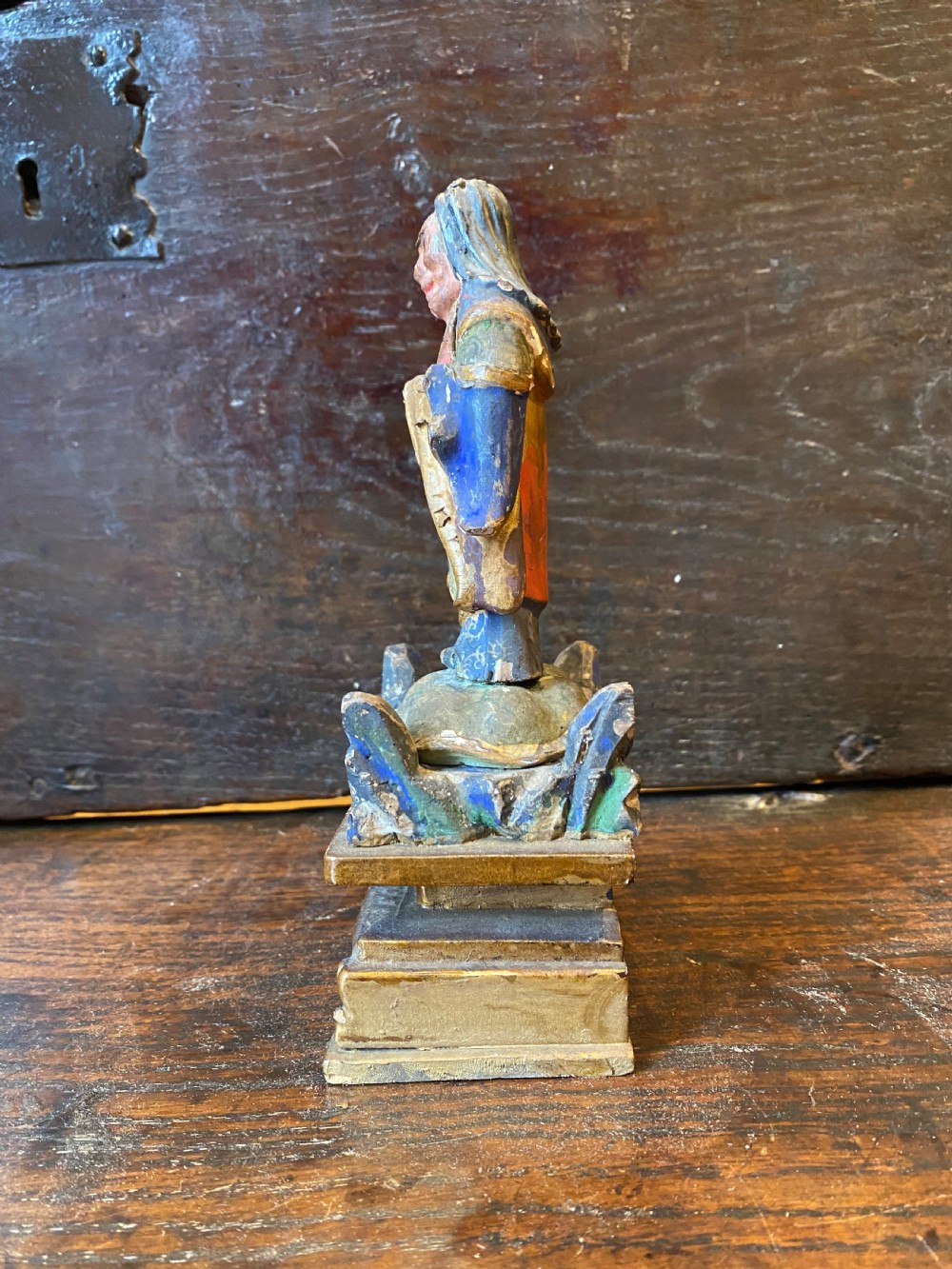 A small hand-carved wooden deity figure with polychrome painted robes and gilt detailing, standing on a carved tiered base. View X