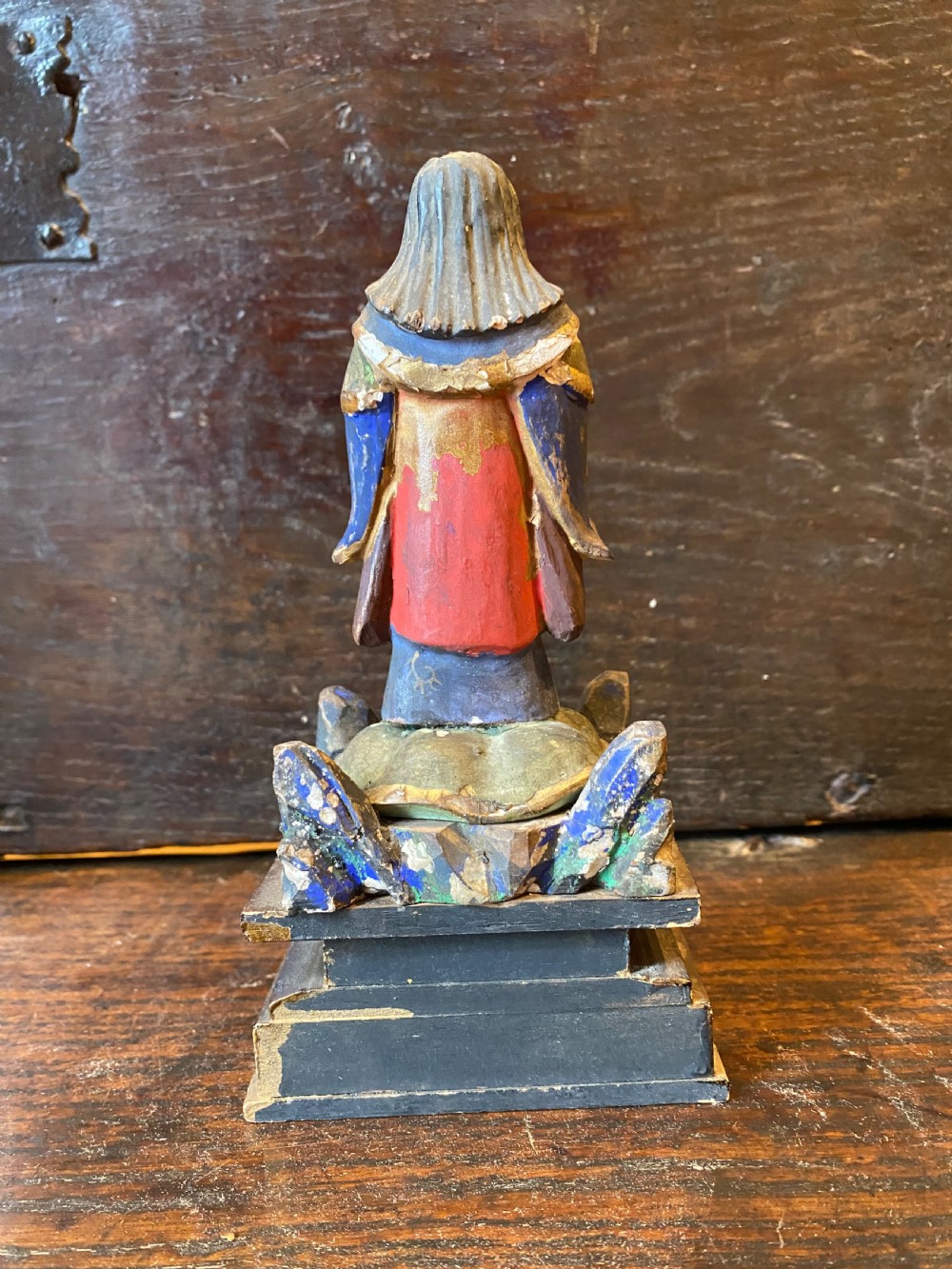 A small hand-carved wooden deity figure with polychrome painted robes and gilt detailing, standing on a carved tiered base. View XI