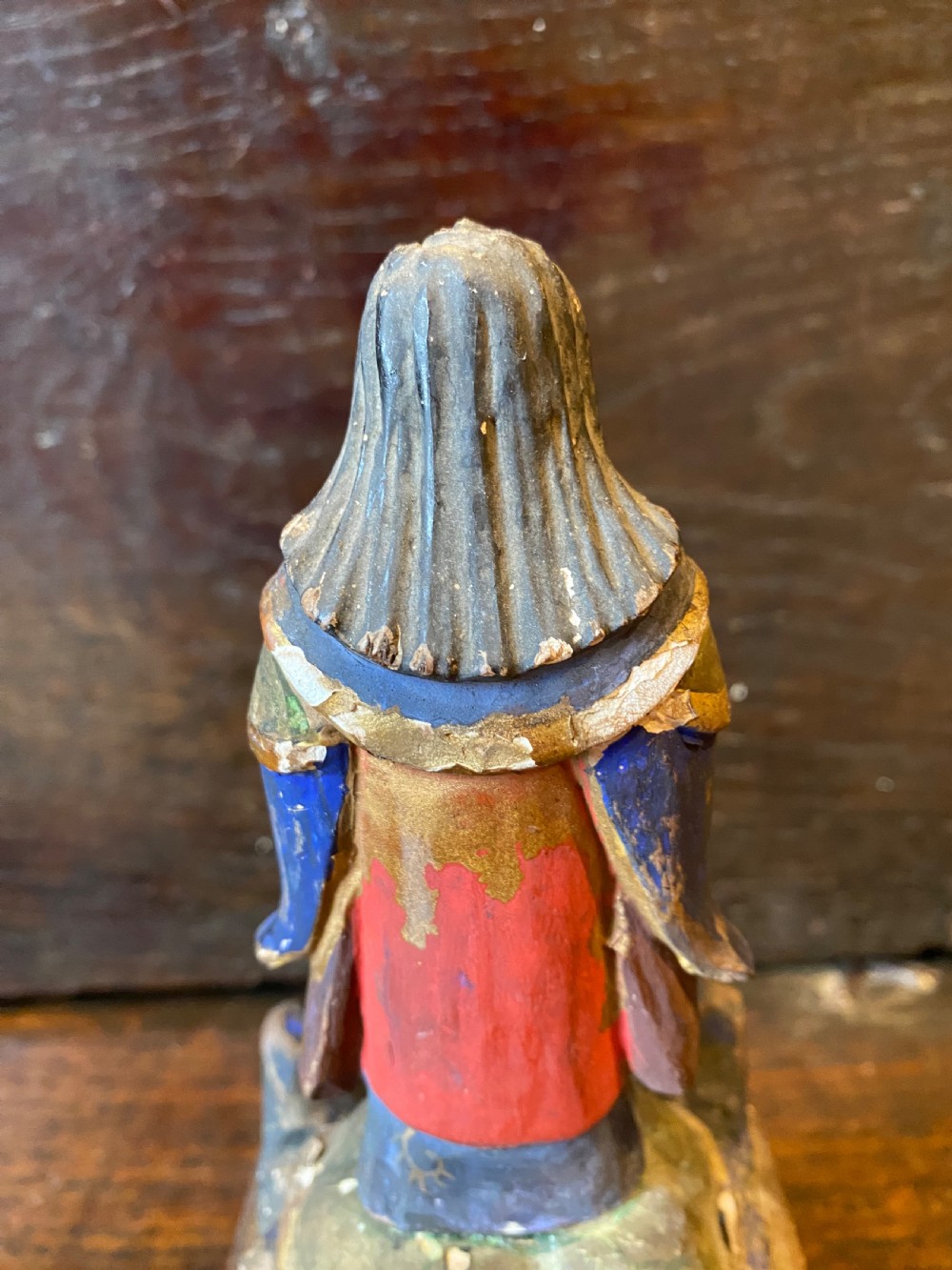 A small hand-carved wooden deity figure with polychrome painted robes and gilt detailing, standing on a carved tiered base. View XII