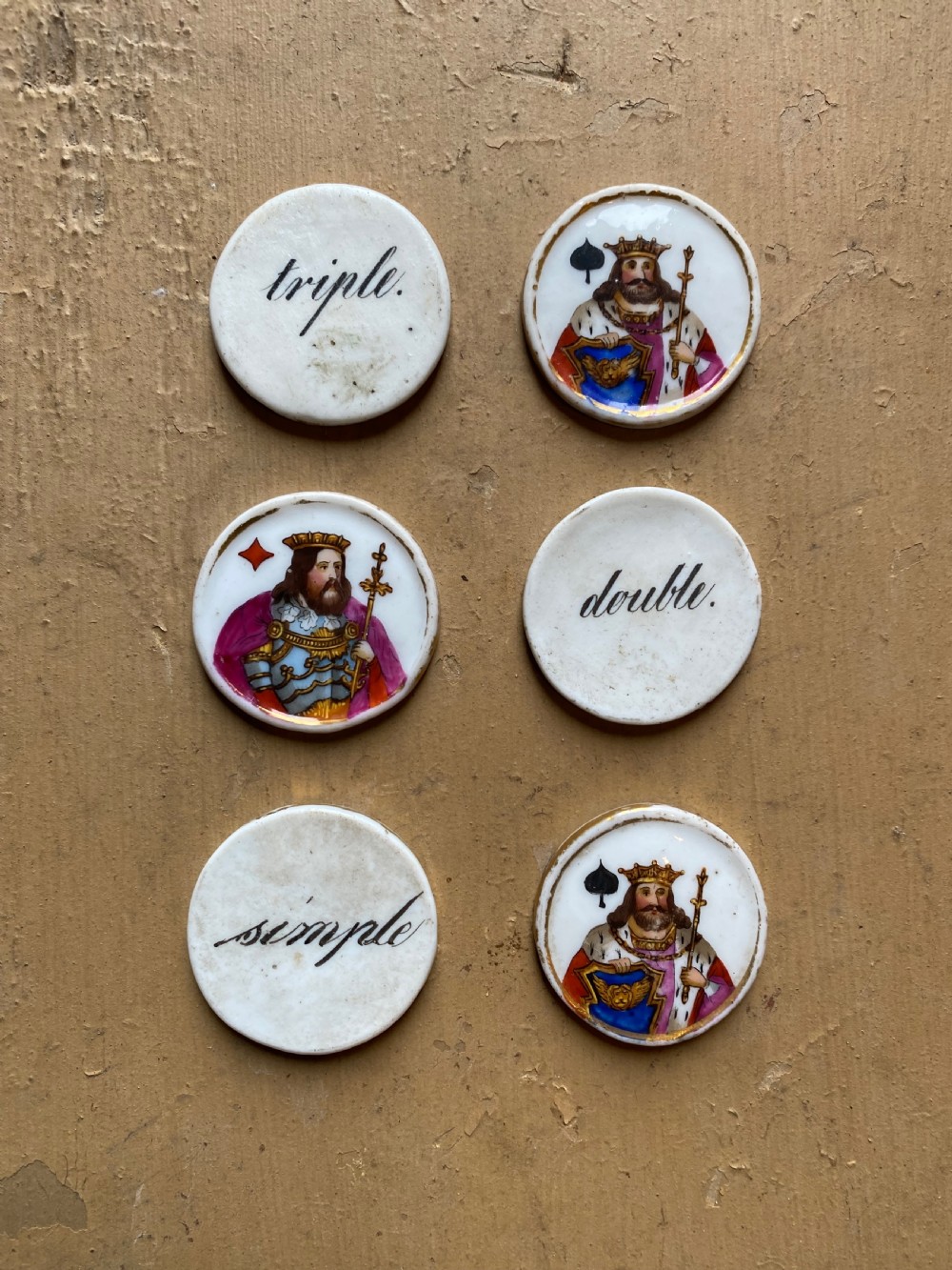 A set of six circular white porcelain gaming tokens decorated with hand-painted playing card king figures and cursive script lettering.