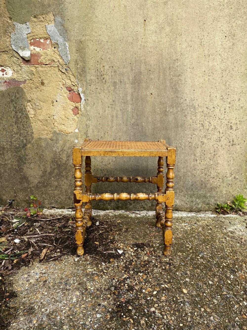 A rectangular wooden stool featuring turned bobbin legs, horizontal stretchers, and a woven cane seat panel with a distressed warm brown finish. View III