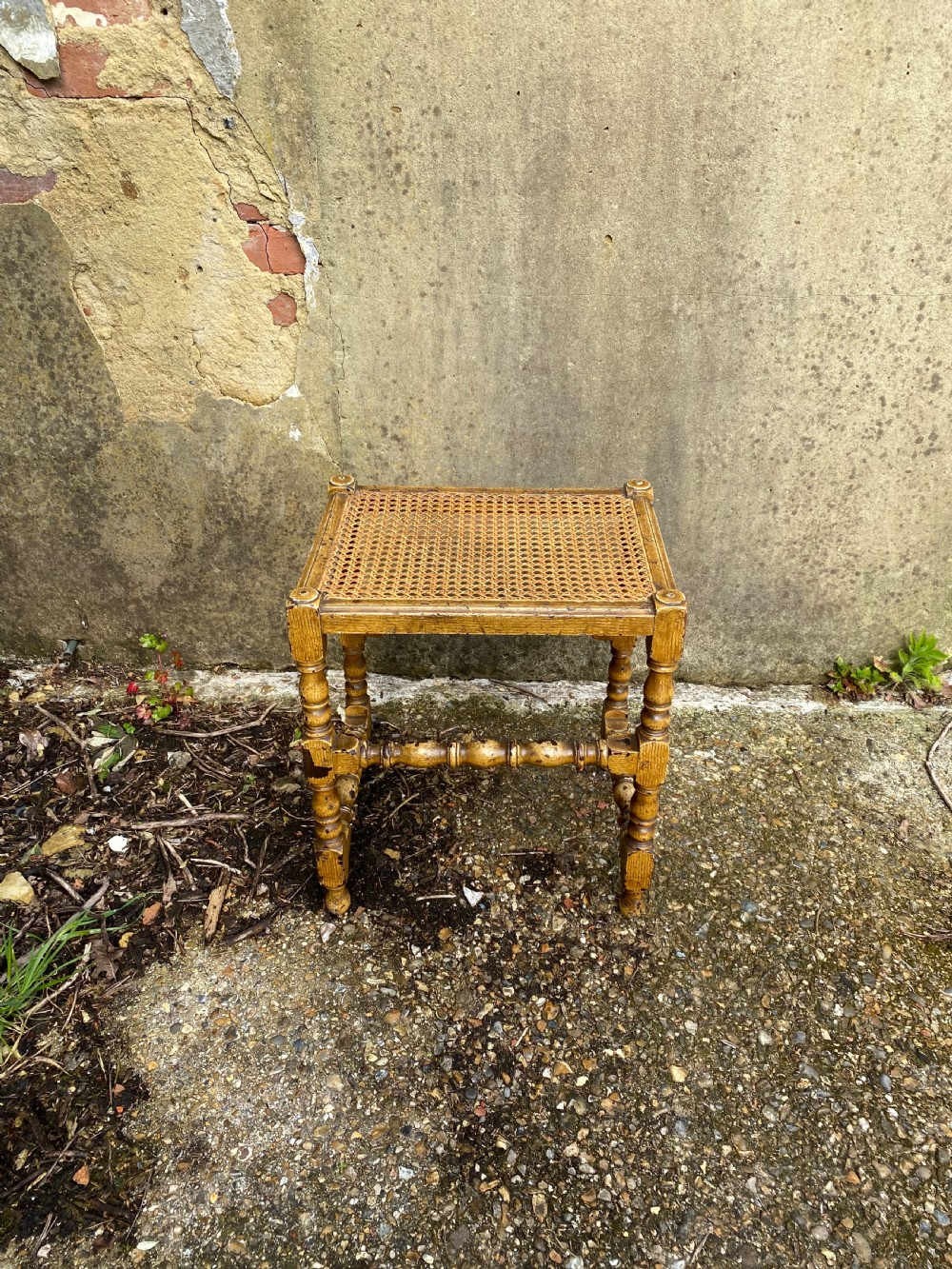 A rectangular wooden stool featuring turned bobbin legs, horizontal stretchers, and a woven cane seat panel with a distressed warm brown finish. View IV