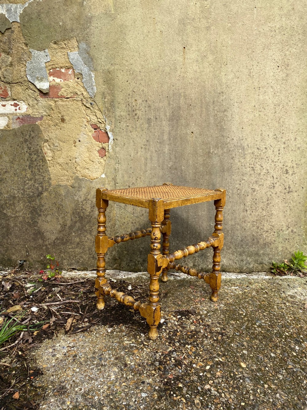 A rectangular wooden stool featuring turned bobbin legs, horizontal stretchers, and a woven cane seat panel with a distressed warm brown finish. View V