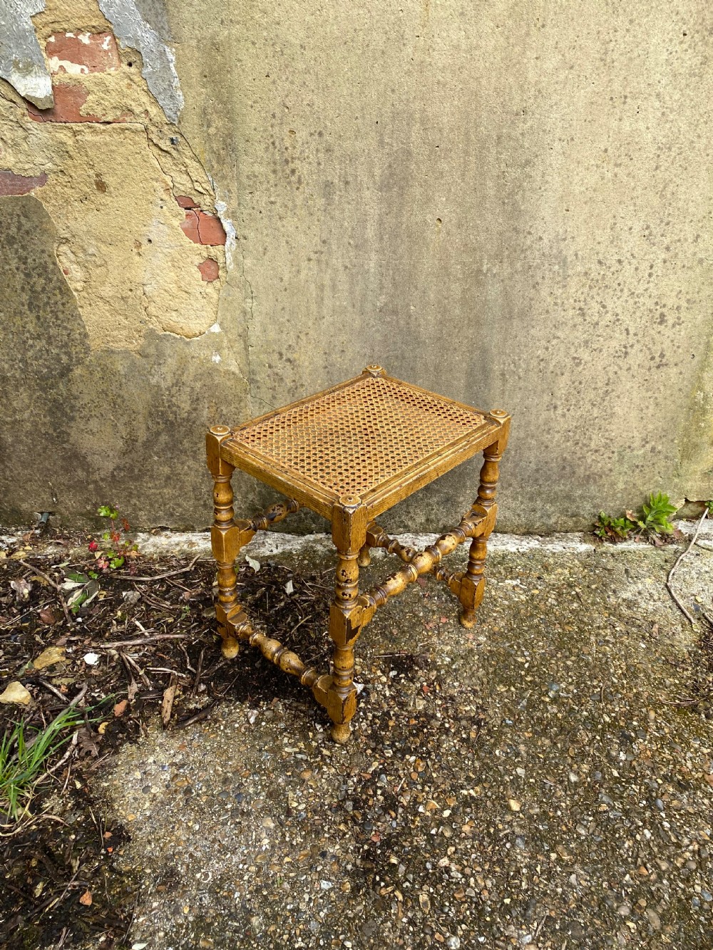 A rectangular wooden stool featuring turned bobbin legs, horizontal stretchers, and a woven cane seat panel with a distressed warm brown finish. View VI