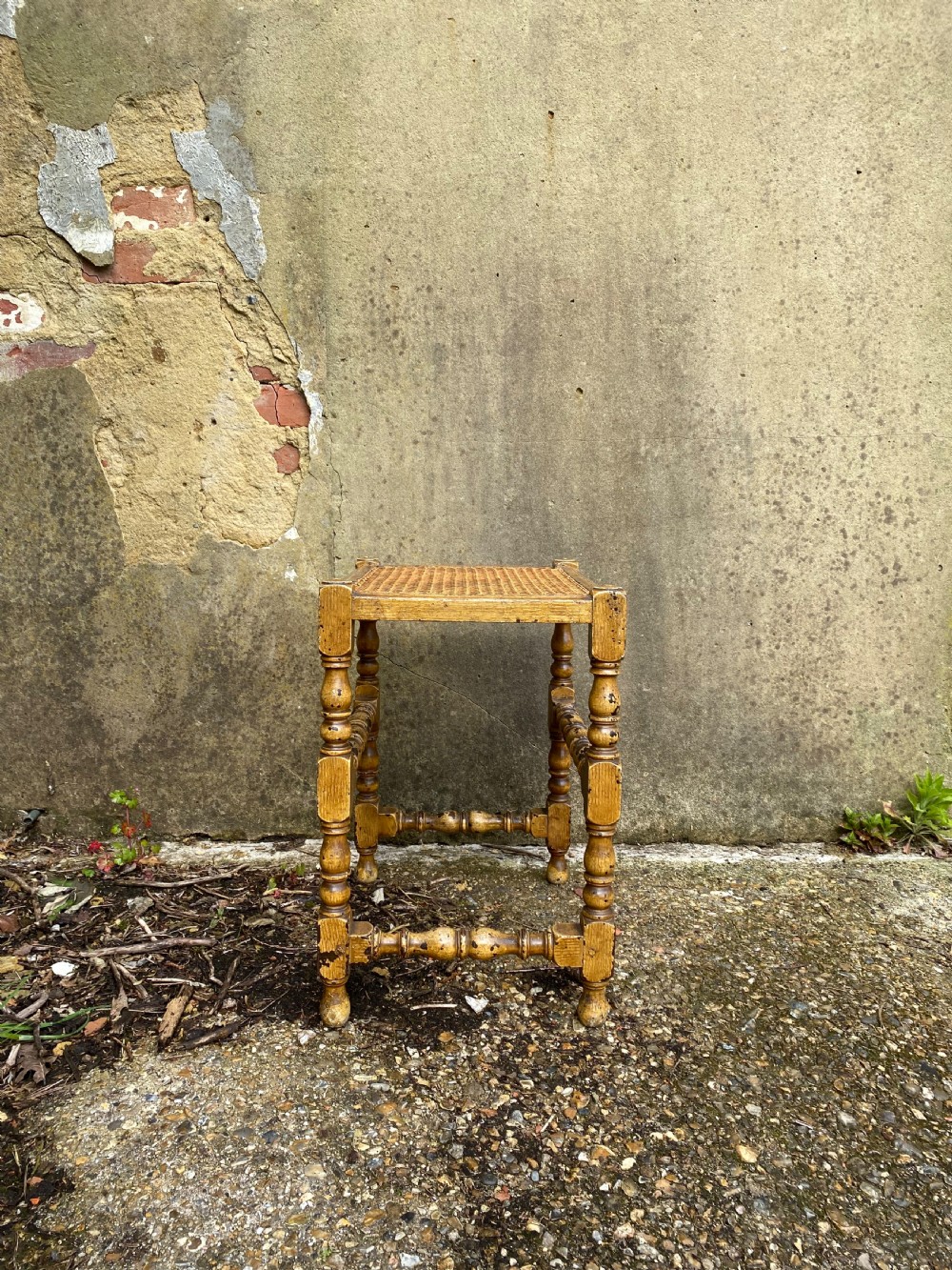 A rectangular wooden stool featuring turned bobbin legs, horizontal stretchers, and a woven cane seat panel with a distressed warm brown finish. View X