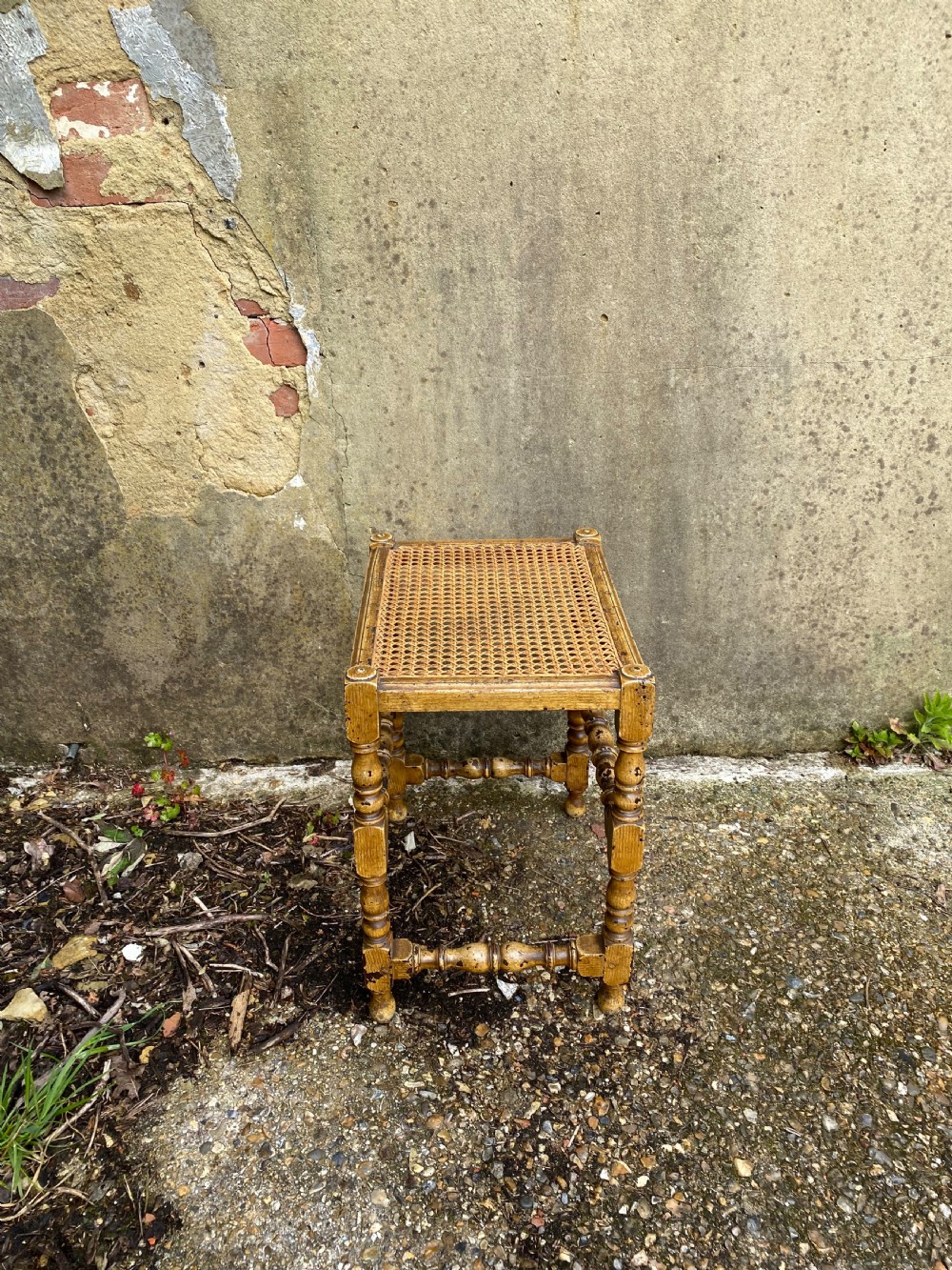 A rectangular wooden stool featuring turned bobbin legs, horizontal stretchers, and a woven cane seat panel with a distressed warm brown finish. View XI