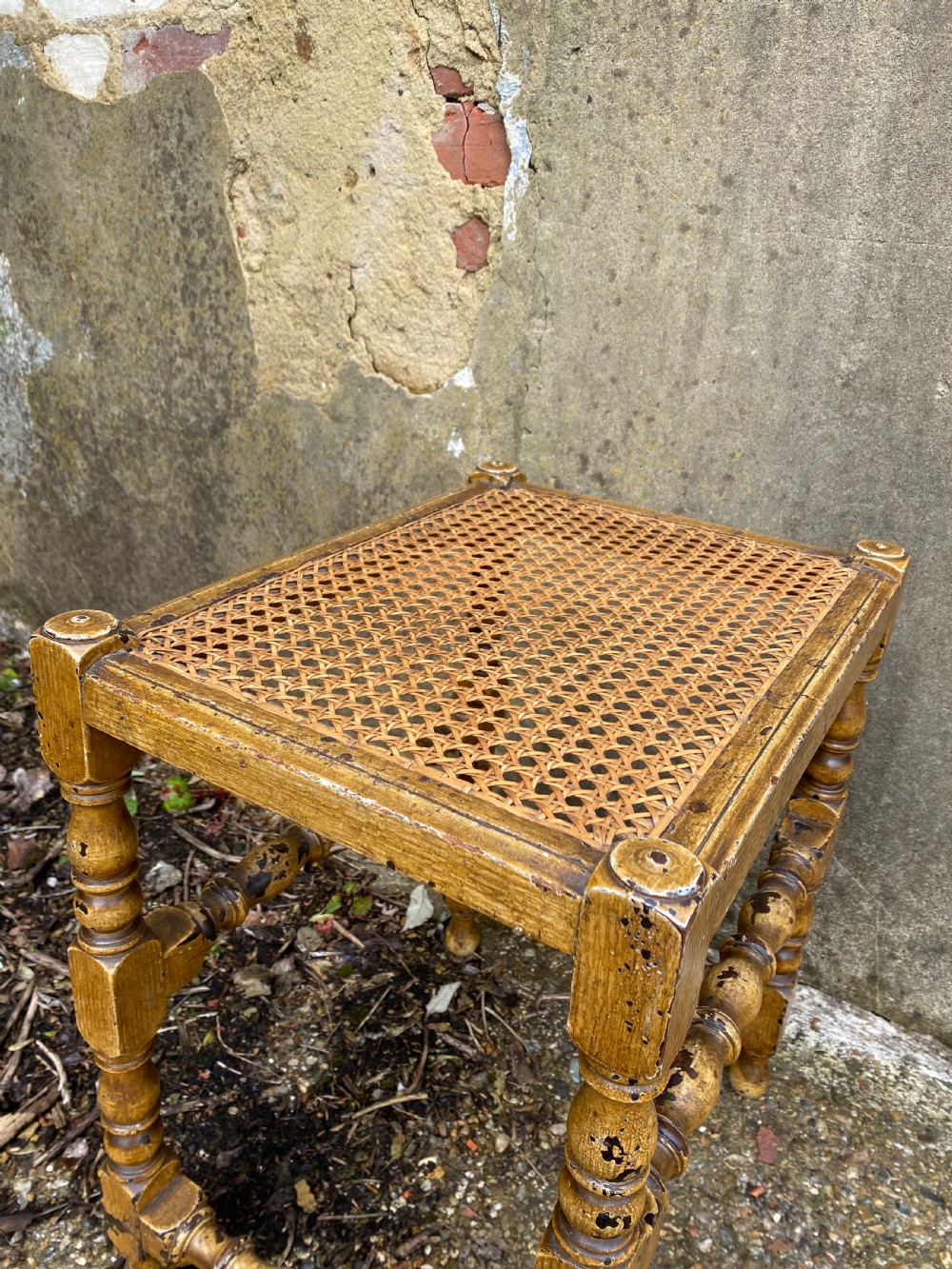 A rectangular wooden stool featuring turned bobbin legs, horizontal stretchers, and a woven cane seat panel with a distressed warm brown finish. View XIII