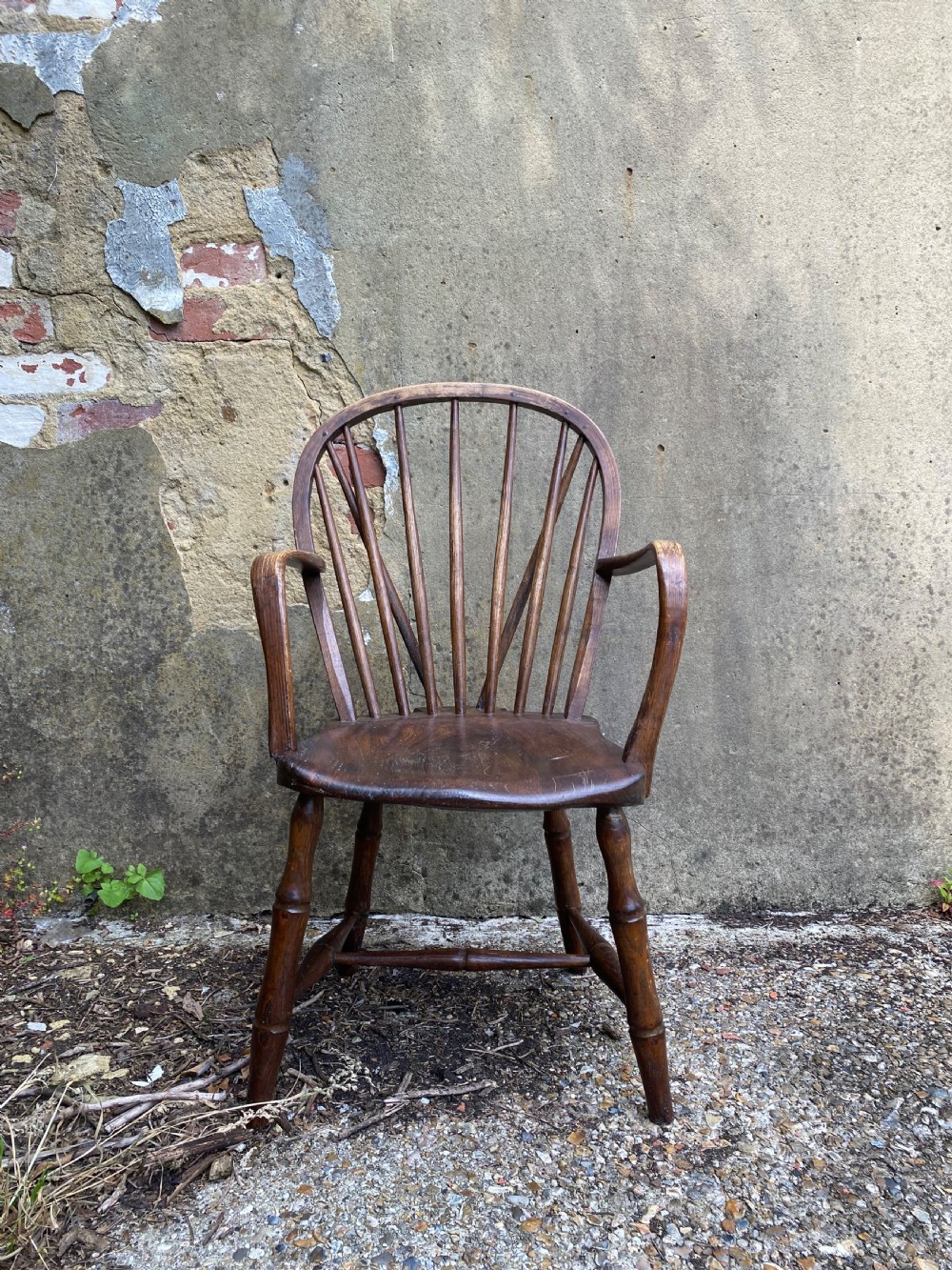 An antique wooden Windsor armchair with a stick-back design and a sculpted wooden seat stands against a weathered wall. View III